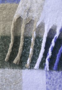 Uldetæppe med frynset kanter. Har et ternet mønster i nuancer af grønt, beige, lyseblå og lilla med en blød, struktureret finish.
