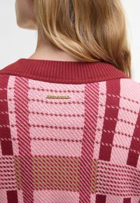 Πλεκτό πουλόβερ με ψηλό γιακά, με καρό σχέδιο σε μπορντό, ροζ και πράσινο χρώμα. Περιλαμβάνει μεταλλική ετικέτα με το όνομα της μάρκας στο πίσω μέρος.