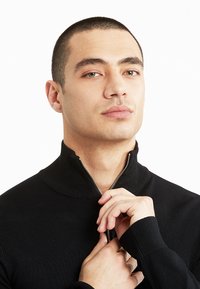Fekete zipzáras pulóver magas gallérral. Az anyag bordázottnak tűnik, a cipzár fémes, ami kiemeli a letisztult designt.