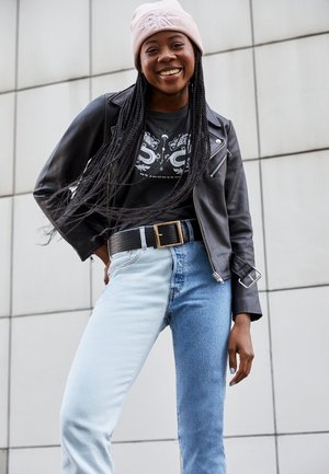 Casaco de cabedal preto estilo biker sobre uma t-shirt gráfica preta, combinado com jeans azul claro e de ganga, e um gorro rosa pálido.
