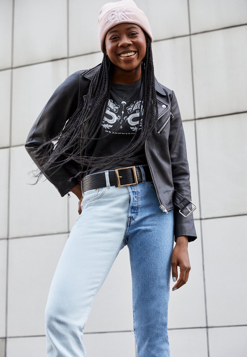 Casaco de cabedal preto estilo biker sobre uma t-shirt gráfica preta, combinado com jeans azul claro e de ganga, e um gorro rosa pálido.