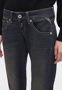 Σκούρο μπλε τζιν από denim με ορατή ραφή, μπροστινό κουμπί, τσέπες και μια μικρή ετικέτα Replay στη δεξιά τσέπη, φορεμένα με μαύρο τοπ.