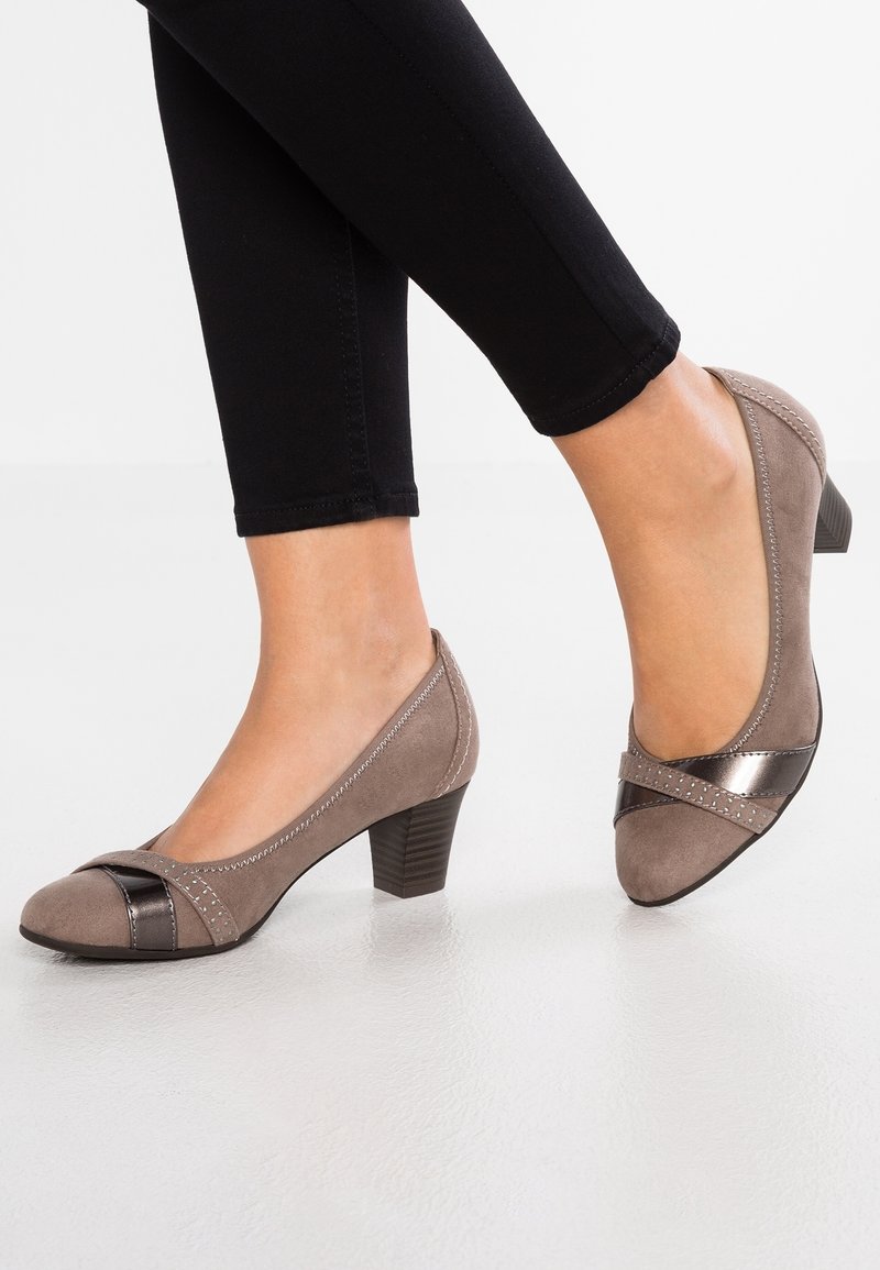 Pieds chaussés de chaussures basses en daim taupe avec des détails métalliques et des coutures sur les brides, assorties à un pantalon noir ajusté.