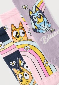 Τρεις πολύχρωμες κάλτσες με παιδικά σκυλίσια πρόσωπα, ουράνια τόξα και λευκά λουλούδια σε φόντα ροζ, μωβ και ναυτικό μπλε.