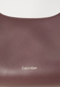Prim-plan al unei genți din piele burgundă, cu un logo auriu "Calvin Klein" pe o suprafață netedă, evidențiind textura și brandingul acesteia.