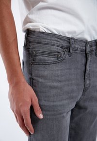 Jeans de denim gris con corte ajustado, que cuentan con remaches metálicos en los bolsillos, una textura sutil y un diseño estándar de cinco bolsillos.