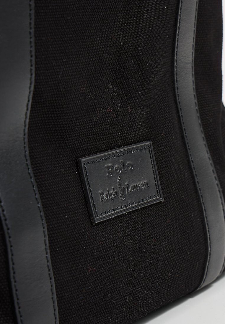 Schwarze Canvas-Tasche mit schwarzen Lederakzenten, ausgestattet mit einem rechteckigen Etikett mit geprägtem Text. Glatte Textur mit matter Oberfläche.