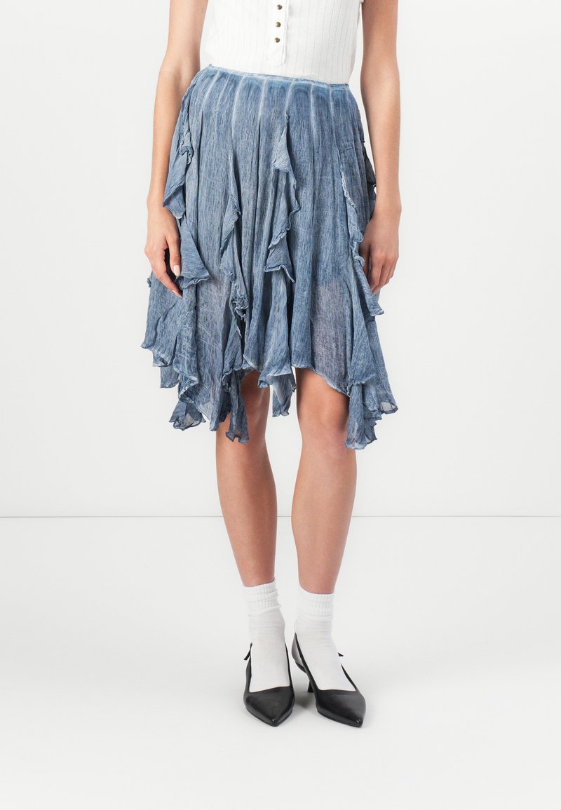 Denim rok met een gerafeld zoom, lichtblauwe kleur, verticale textuur en een asymmetrische snit. Gepaarde met zwarte puntige schoenen en witte sokken.