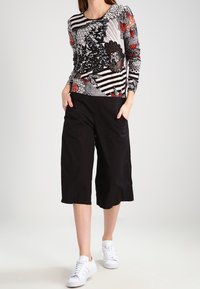 Kvinna i ett mönstrat långärmat plagg, svarta vida capribyxor och vita sneakers, står med händerna i fickorna.