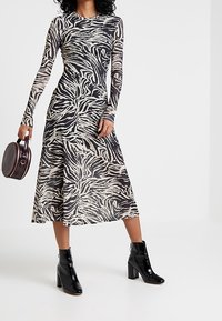 Långärmad klänning i midilängd med svart och beige zebra-mönster, ihop med svarta lackstövlar och en rund, mörk handväska.