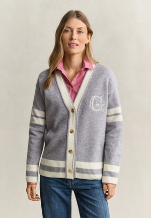 Femme portant un cardigan gris avec des rayures crème et des boutons dorés sur une chemise rose à col et un jean bleu, debout devant un fond uni.