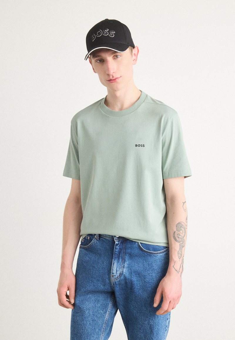 Lys grønn bomulls T-skjorte med trykt "BOSS"-logo, kombinert med blå denimjeans. Modellen har på seg en svart caps med hvit stikking.