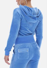 Blue velour tracksuit featuring a hooded top and elastic waistband. Pockets on pants have snap closures, with a soft, smooth texture.