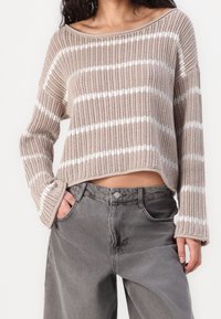 Helles, beiges Strickpullover mit weißen horizontalen Streifen, weitem Ausschnitt, lockerer Passform und langen Ärmeln. Kombiniert mit grauen Denim-Hosen.
