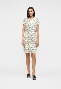 Γυναίκα που φοράει κρεμ φόρεμα μέχρι το γόνατο με κοντά μανίκια και μαύρα αφηρημένα γεωμετρικά σχέδια, καθώς και μαύρα ανοιχτά σανδάλια, στέκεται σε λευκό φόντο.