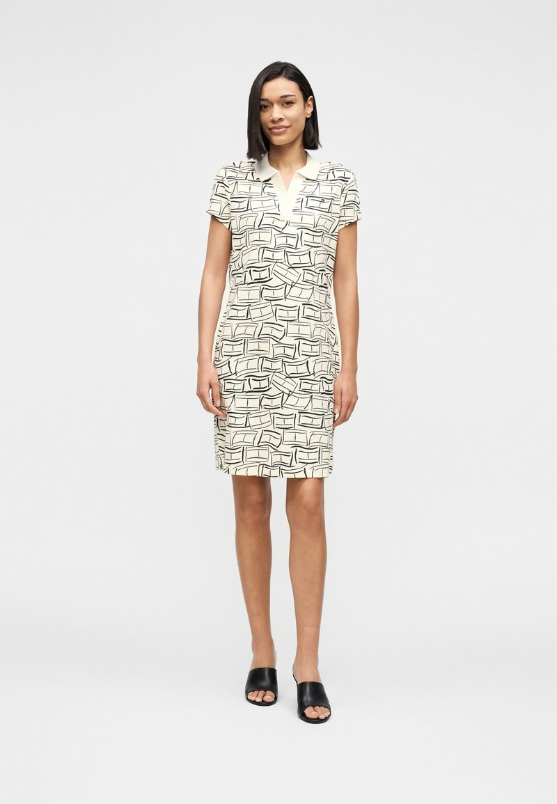 Γυναίκα που φοράει κρεμ φόρεμα μέχρι το γόνατο με κοντά μανίκια και μαύρα αφηρημένα γεωμετρικά σχέδια, καθώς και μαύρα ανοιχτά σανδάλια, στέκεται σε λευκό φόντο.