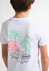 Bílý bavlněný tričko s grafikou znázorňující zelenou plážovou scénu, palmami, záchrannou věží v oranžové barvě a siluetou černého auta.