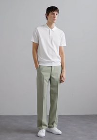 Hvit kortermet polo skjorte, lysegrønne skreddersydde bukser og hvite sneakers, står mot en nøytral bakgrunn. Minimalistisk design.