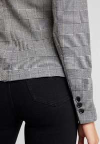Schwarze und weiße Karoblazerärmel mit drei schwarzen Knöpfen am Bündchen; kombiniert mit eng anliegenden schwarzen Jeans. Die Textur wirkt glatt.