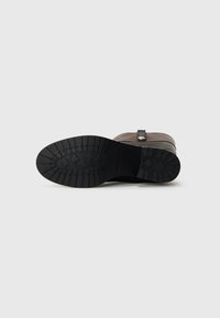 Bottine en cuir marron avec une semelle en caoutchouc noire, dotée d'un design de semelle texturée et d'une sangle latérale avec un accent métallique.