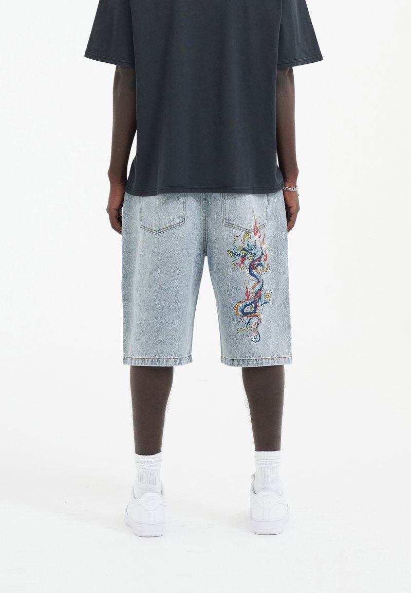 Pantalones cortos de mezclilla azul claro con intrincados bordados de dragón en colores vibrantes en el lado; combinados con una camiseta oscura holgada y zapatillas blancas.