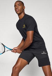 Black athletic shirt with a yellow logo on chest, paired with black shorts. Model grips a blue and black tennis racket.