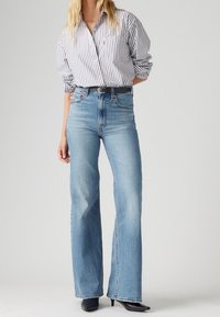 Persona con unos vaqueros de tiro alto en azul claro acampanados, un cinturón negro, unos zapatos de tacón negro de punta y una blusa blanca con rayas verticales negras.