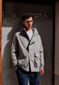 Mladý muž s tmavými kudrnatými vlasy, oblečený v světlé šedé bundě s kapucí a tmavě modrých kalhotách, stojící před zastíněnými dřevěnými dveřmi.