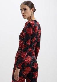 Top a maniche lunghe in velluto nero con un ampio motivo di rose rosse, design aderente e texture liscia. Modello mostrato di lato.