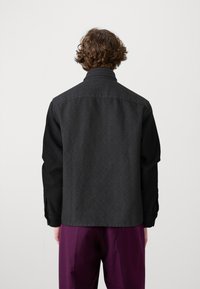 Czarna kurtka w wzory z kołnierzem koszulowym, długimi rękawami i prostym krojem. Noszona z fioletowymi spodniami, pokazująca fakturowany tył.