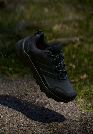 Czarny but trekkingowy Adidas Terrex zawieszony w powietrzu nad żwirową ścieżką z rozmytym zielonym trawiastym tłem.