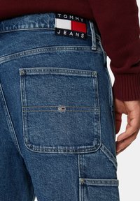 Jeans in denim di colore blu scuro con una tasca posteriore che presenta un piccolo patch logo in rosso, bianco e blu, e dettagli di cucitura visibili.