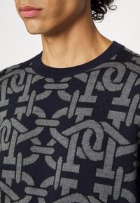 Maglione blu scuro con un motivo geometrico grigio. Il tessuto sembra lavorato a maglia, con un colletto a costine e disegni intricati e intrecciati.