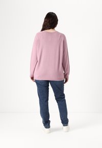 Ανοιχτό ροζ πλεκτό πουλόβερ με στρογγυλή λαιμόκοψη και μακριά μανίκια, σε συνδυασμό με μπλε τζιν παντελόνι και λευκά αθλητικά παπούτσια, φαίνεται από πίσω.