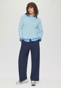 Un pull côtelé bleu clair superposé à une chemise à rayures bleues, associé à un pantalon ample bleu marine et des baskets argentées.