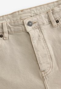 Beige relaxed fit denim pants with a top button and multiple pockets. Visible stitching in a lighter thread, with textured fabric.
