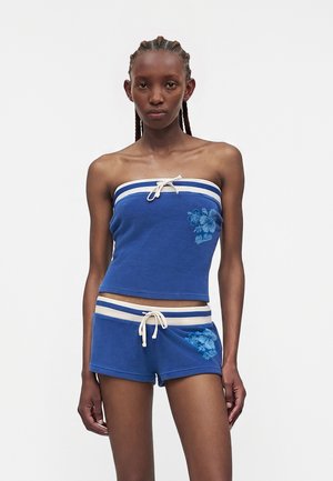 Jeune femme portant un haut bustier bleu et un short assorti avec des rayures blanches et une broderie florale, debout devant un fond uni.