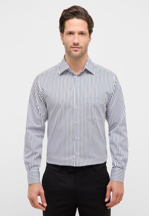 Mand iført en hvid langærmet skjorte med sorte lodrette striber og sorte bukser, stående mod en ensartet lys baggrund.