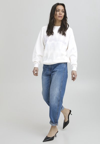 Sudadera blanca con el texto "BALL" en diferentes groserías, combinada con jeans baggy azul claro y tacones negros de punta. Texturas suaves en todo.
