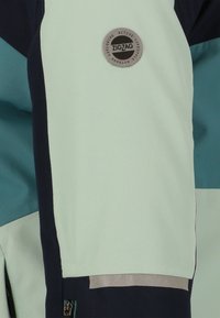 Giacca verde chiaro e blu navy con un tessuto testurizzato, accessori in metallo argentato e un logo circolare che recita "ZIGZAG" nella zona del petto sinistro.