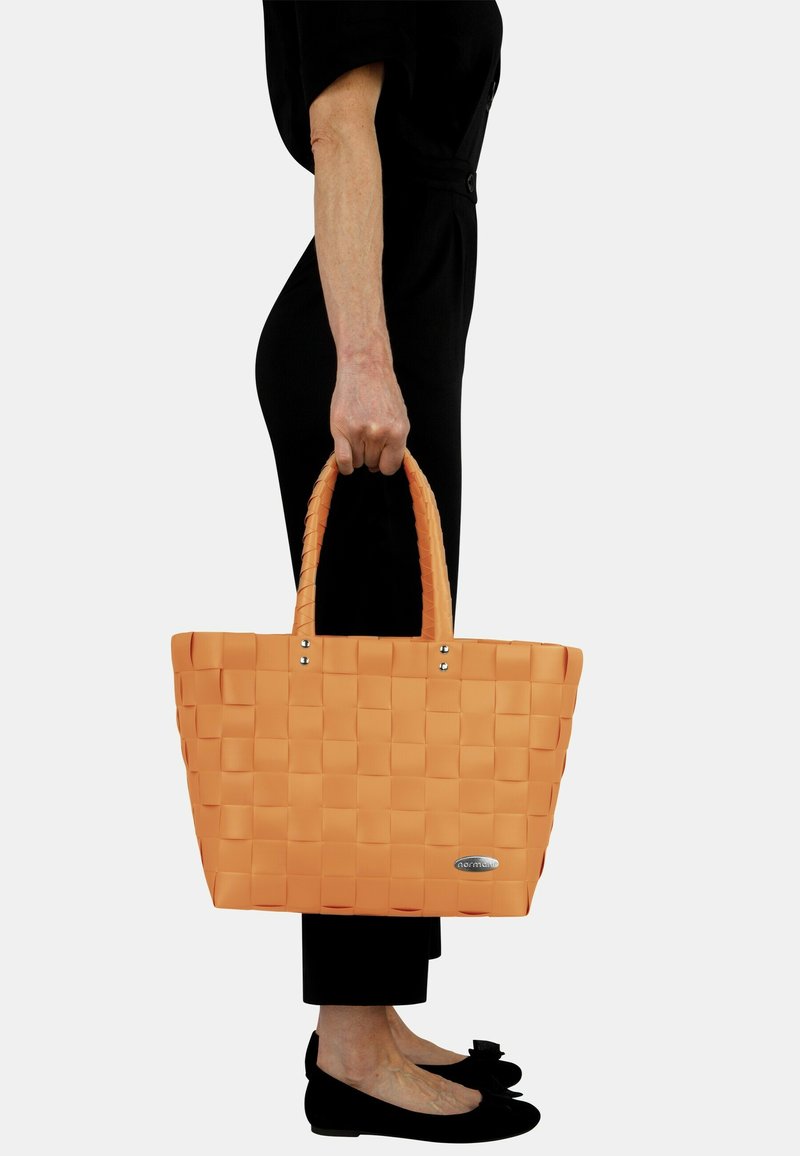 Orange geflochtener Tragetasche mit zwei kurzen Griffen, ausgestattet mit silbernem Zubehör und einem glänzenden Logo. Glatte Textur mit rechteckiger Form.