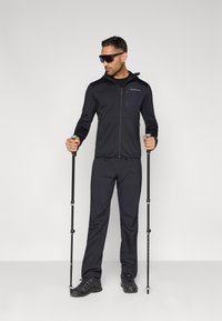 Czarna sportowa kurtka z zamykanymi kieszeniami, w połączeniu z dopasowanymi spodniami i ciemnym obuwiem. Użytkownik trzyma kijki trekkingowe, ma na sobie okulary przeciwsłoneczne.