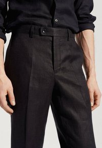 Man wearing black tailored trousers and a black long-sleeve shirt with rolled-up sleeves, hands resting by sides.