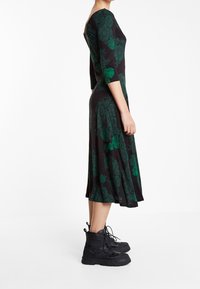 Robe florale noire avec des motifs verts, manches trois-quarts et ourlet évasé, portée avec des bottines noires. Texture de tissu lisse.