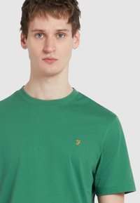 T-shirt verde in cotone con collo rotondo, maniche corte e piccolo logo arancione sul petto. Tessuto liscio senza motivi visibili.