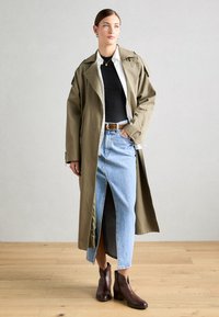 Olivengrønn trenchcoat over en svart topp, kombinert med en lys blå denimskjørt med en frontslit, og brune ankelstøvler.