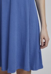 Ein knielanges, strukturiertes blaues Kleid mit leicht ausgestellter Form, das aus einem weichen, zerknitterten Stoff gefertigt ist und einen schlichten Saum aufweist.