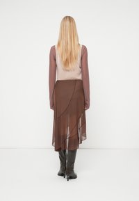 Καφέ διαφανής φούστα με ασύμμετρο στρίφωμα. Το μοντέλο φοράει μια εφαρμοστή, μακρυμάνικη μπλούζα σε πιο ανοιχτό τόνο, συνδυασμένη με μαύρες μποτες έως το γόνατο.