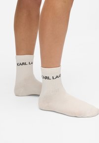 Kreminės medvilninės kojinės su raukšlėmis, viršuje esančiu juodu tekstu "KARL LAGERFELD". Skaidri tekstūra ir vidutinio ilgio dizainas iki blauzdos.