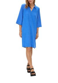 Robe en lin bleue, longueur genoux, avec un décolleté en V, des manches trois-quarts et des fentes latérales. Chaussures noires à plateforme avec des brides.
